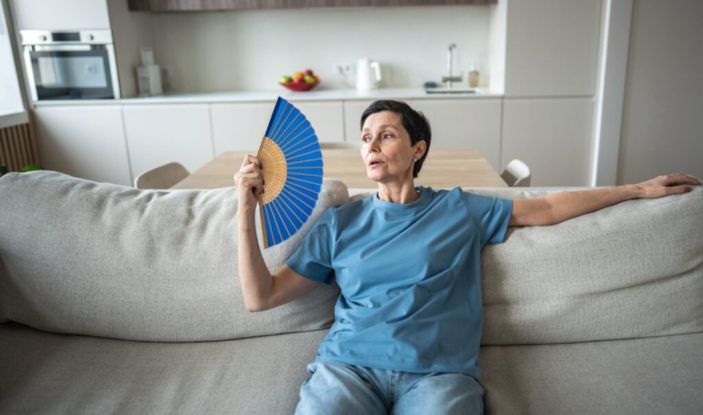 Woman sitting on a couch fanning herself to cool down in a warm home.